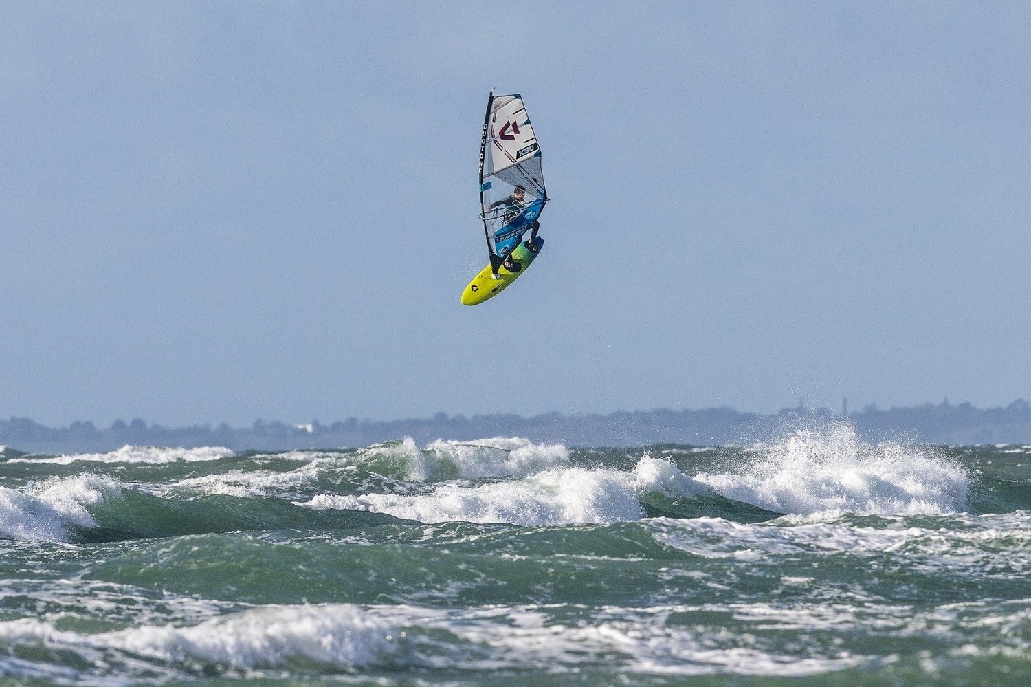 A windsurfer launching a high jump above a green-grey wave under blue sky